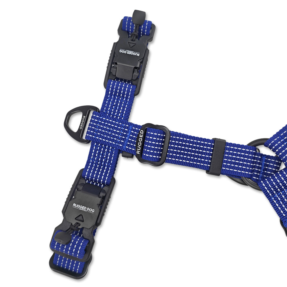 Close-up top view of Navy Rugged Dog Company no-pull harness showing dual magnetic FIDLOCK V-Buckles, aircraft-grade aluminum back D-ring for standard leash walking, Rugged-branded metal adjustment sliders and high-visibility reflective polyester webbing